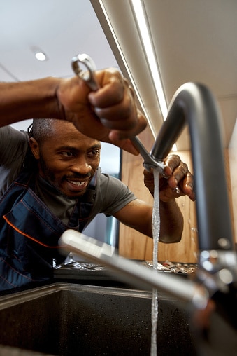 Low angle view African American plumber in overalls fixing water tap with leaking water flow using spanner. Black modern sink with dark faucet. Authenticity in business concept.